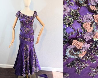 May include: A purple, floor-length gown with a fitted bodice and a flowing skirt. The bodice is covered in intricate beading and embroidery, featuring floral motifs in shades of pink and brown. The gown is displayed on a mannequin and has a label that reads "Fezandi Beverly Hills".