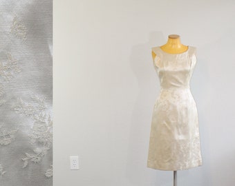 May include: A white sleeveless dress with a floral pattern on a mannequin. The dress is on a stand with a silver base. The dress is in a room with a wood floor.