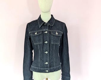 May include: A dark indigo denim jacket featuring a classic collar, long sleeves with buttoned cuffs, and a front button closure. The garment includes two chest pockets with buttoned flaps and visible white contrast stitching throughout the design.