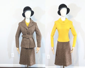 May include: A brown tweed skirt suit with a matching jacket and skirt. The jacket has a belt and the skirt is knee-length. The suit is displayed on a mannequin wearing a yellow sweater and a black hat.
