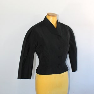 May include: A black cropped jacket with a collar and button closure. The jacket is on a mannequin with a yellow skirt.