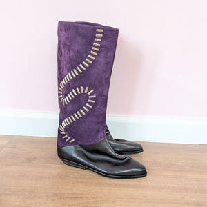 May include: A knee-high boot featuring a deep purple suede upper with a decorative, winding light-colored stitched pattern along the side. The lower portion of the boot is constructed from smooth black leather, featuring a flat sole and a rounded toe.