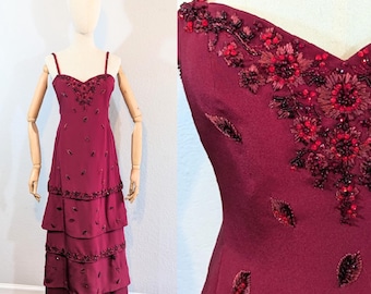 May include: A burgundy floor-length gown with a sweetheart neckline and spaghetti straps. The dress is adorned with red and black beading and sequins, creating a floral pattern. The skirt is tiered and flows gracefully.