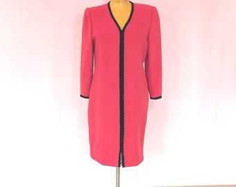 May include: A vibrant coral-pink knee-length dress featuring long sleeves, a V-neckline, and a central vertical black trim detail. The garment includes black piping along the neckline and sleeve cuffs, creating a structured and clean silhouette.