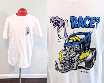 May include: White t-shirt featuring a cartoon hot rod design with the text "RACE?" on the back. The front of the shirt has a small graphic. The design includes a purple monster hand, a blue car, and yellow flames. The shirt is displayed on a mannequin.