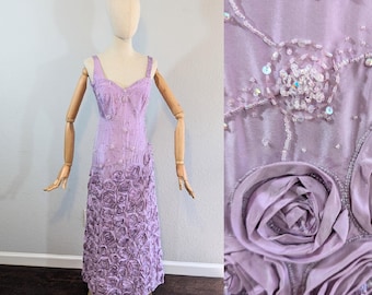 May include: A lavender formal dress with a sweetheart neckline and spaghetti straps. The bodice is embellished with beads, and the skirt features a cascade of fabric roses. The dress is displayed on a mannequin.