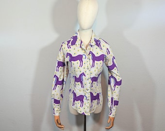 May include: A button-down shirt with a white background and a repeating pattern of purple horses. The shirt has long sleeves and a collared neckline. Small yellow dots are scattered across the white background.