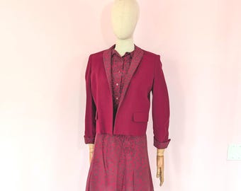 May include: A deep magenta cropped blazer with long sleeves and a shawl collar, worn over a matching button-down shirt and a pleated skirt. The shirt and skirt feature a dark grey botanical pattern, while the blazer cuffs show the same print.
