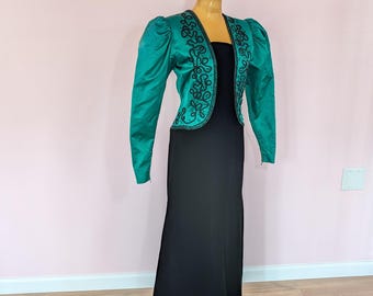 May include: A floor-length black dress paired with a cropped emerald green jacket. The jacket features long puff sleeves and intricate black corded embroidery along the front edges. The dress has a straight silhouette and a simple, sleeveless neckline.