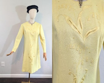 May include: A pale yellow dress with long sleeves and a round neckline. The dress is embroidered with a floral design in a darker yellow thread. The dress is on a mannequin.
