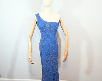 May include: A blue, one-shoulder evening gown with intricate beaded detailing. The dress is displayed on a mannequin with wooden arms and a tiara. The gown's design features a fitted silhouette that flows to the floor.