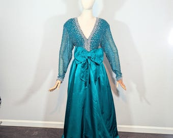 May include: A teal-colored formal gown with a sequined bodice and long sleeves. The dress features a large bow at the waist and a flowing skirt. A silver tiara sits atop the mannequin's head. The dress is displayed on a mannequin.
