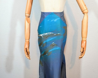 May include: A blue and teal skirt with a unique design. The skirt features a print of a seascape with mountains and waves. The skirt is displayed on a mannequin. The skirt has a high waist and a flared hem.