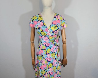 May include: A wrap dress with a vibrant floral pattern in pink, blue, and yellow on a navy blue background. The dress features short sleeves and a V-neckline. The dress is displayed on a mannequin.