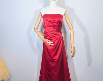 May include: A strapless, floor-length red satin gown displayed on a mannequin. The dress features a fitted bodice with a ruched detail on the side and a flowing skirt. The mannequin has wooden arms and a neutral-colored head.