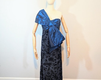 May include: A one-shoulder, floor-length evening gown. The bodice features a large, draped bow in a vibrant blue. The skirt is a dark navy with a subtle floral pattern. A silver tiara sits atop the mannequin's head.