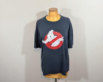May include: A dark blue t-shirt featuring the Ghostbusters logo. The logo is a white ghost inside a red circle with a red line through it. The shirt is displayed on a mannequin.