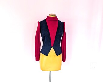 May include: A long-sleeved red turtleneck top layered under a dark navy blue vest. The vest features a deep V-neckline, structured shoulders, and a pointed hemline that meets at the center. The garment is displayed on a mannequin torso.