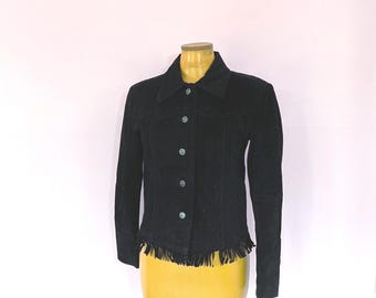 May include: A black suede jacket featuring a pointed collar, long sleeves, and a front button closure. The garment is detailed with a horizontal stitched seam across the chest and a fringe trim along the bottom hemline, providing a textured finish.