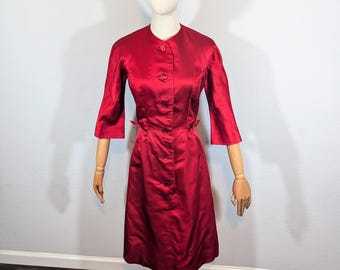May include: A vintage red satin dress with a button-up front and three-quarter sleeves. The dress is knee-length and features a fitted waist with small belt loops. The dress is displayed on a mannequin with wooden arms and a gold-colored base.