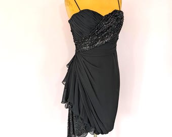 May include: A black sleeveless dress featuring a sweetheart neckline and thin spaghetti straps. The bodice is adorned with a diagonal band of black sequins, while the skirt displays draped fabric layers and an asymmetrical hemline with sequin detail.