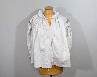 May include: Cream-colored long-sleeved blouse with a ruffled collar and blue embroidered details. The blouse is displayed on a mannequin. The fabric appears to be linen or a similar lightweight material. The blouse has a loose fit and a V-neck.