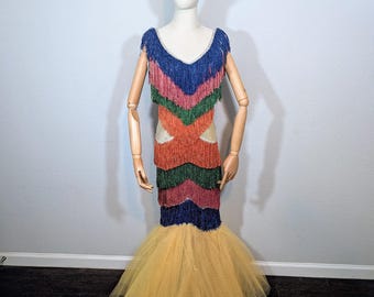 May include: A colorful, fringed dress with a V-neckline and a mermaid-style skirt. The dress features layers of blue, pink, green, orange, and burgundy fringe. The skirt is made of yellow tulle. The dress is displayed on a mannequin.