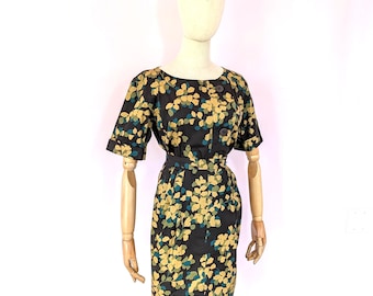 May include: A black dress featuring a repeating pattern of yellow and teal leaves. The garment has short sleeves with cuffs, a round neckline, a fitted waist with a matching belt, and a button-down front closure with two visible dark buttons.