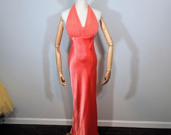 May include: A coral-colored halter neck gown on a mannequin. The dress has a flowing, floor-length design with a fitted bodice and a flared skirt. The fabric appears to have a satin-like sheen, reflecting light. The mannequin has wooden arms and a neutral-colored head.