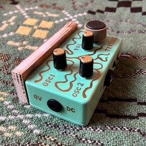 May include: A mint green electronic audio effects pedal with black knobs and gold accents. The pedal has the text "OSCI", "VOL", and "TEXTURE" in a brown font. A wooden frame is attached to the side of the pedal.