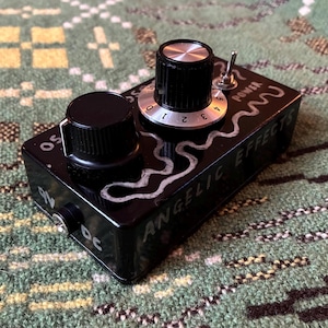 May include: Black guitar effects pedal with two black knobs, a silver toggle switch, and the text "Angelic Effects". The pedal has a silver dial with numbers 0-5. The pedal is on a green patterned surface.