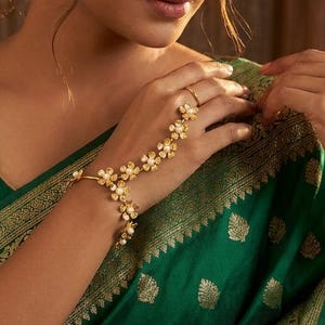 May include: A gold hand harness featuring a delicate chain design adorned with small, five-petaled flower motifs that connect a wrist bracelet to a single finger ring, worn with a deep emerald green fabric featuring gold woven patterns.