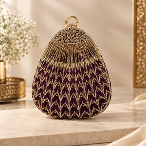 May include: A teardrop-shaped purple clutch bag featuring intricate gold embroidery and beadwork in a chevron pattern. The top of the bag is adorned with a dense cluster of gold beads and crystals, finished with a circular gold metal ring handle.
