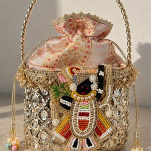 May include: A cylindrical metallic gold handbag with a mirrored finish and a twisted gold handle. The top features a drawstring closure made of patterned fabric. A large, colorful beaded applique depicting a stylized figure is attached to the front.