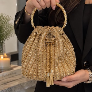May include: A gold bucket-style handbag featuring an intricate exterior covered in shimmering crystals and beads. The bag has a circular gold metal handle and a drawstring closure with two long, dangling gold chain tassels at the front center.