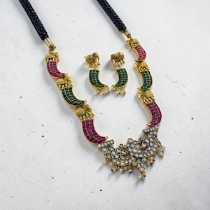 May include: An ornate necklace and earring set featuring gold-toned elephant motifs and curved, jewel-toned accents in green and magenta. The necklace has a black cord and a detailed pendant with white gemstones.