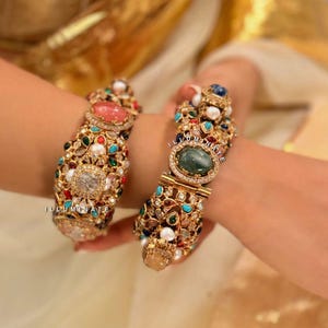 May include: Two ornate gold bracelets encrusted with a variety of colorful gemstones and pearls. The bracelets feature large oval and rectangular stones in shades of pink, green, and blue. The word "ILLUMINATE" is visible on the bracelets.