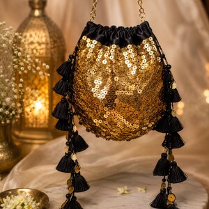 May include: A drawstring pouch bag covered in shimmering gold sequins with a black satin top. The bag features long, dangling black tassels accented with small gold circular charms and is suspended by a thin gold metal chain strap.