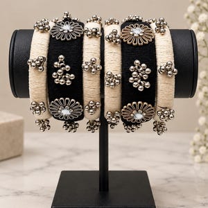 May include: A set of six bangles arranged on a display stand. The bangles alternate between cream-coloured thread-wrapped bands and black thread-wrapped bands. The black bangles feature silver-toned floral medallions and clusters of small silver beads.