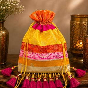 May include: A drawstring pouch featuring horizontal bands of orange, yellow, hot pink, and gold fabric with a floral pattern. The bag is cinched at the top with yellow cords and decorated with a row of dangling hot pink tassels along the bottom edge.