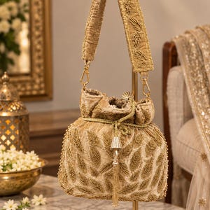 May include: A gold velvet bucket bag featuring intricate beadwork in a leaf pattern. The bag includes a matching shoulder strap with beaded leaf details, a drawstring closure with a metallic tassel, and gold-toned hardware clips on both sides.