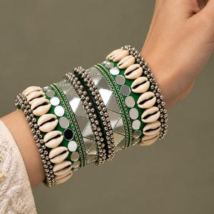 May include: A set of wide bangles featuring a vibrant green fabric base adorned with rows of cream-colored cowrie shells, small metallic silver beads, and triangular mirror accents arranged in a geometric pattern across the surface.