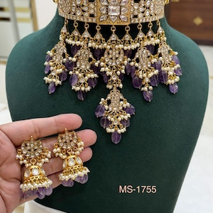 May include: A gold-toned necklace and earring set with intricate detailing. The necklace features a wide choker design with dangling pendants adorned with purple gemstones and small white pearls. The earrings mirror the necklace's design. The text "MS-1755" is visible.