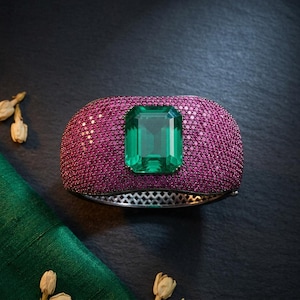 May include: A wide, ornate bracelet featuring a large, emerald-cut green gemstone at the center. The bracelet's band is covered in small, closely set pink gemstones. The setting appears to be silver or a light-colored metal.