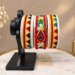 May include: Three bangles displayed on a black stand. The two outer bangles feature a white base with a repeating red, yellow, and blue triangular pattern. The center bangle has a black base with colorful embroidered floral motifs and mirrors.