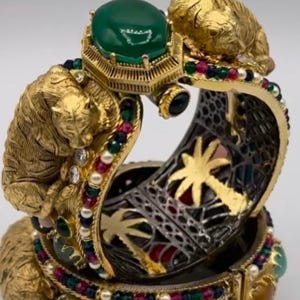 May include: Ornate gold-toned bracelet with intricate details. Features a large green gemstone, small gemstones in various colors, and two sculpted tiger figures. The bracelet has a detailed openwork design with gold palm tree accents.