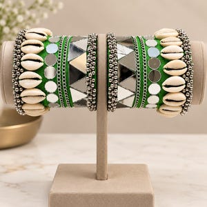 May include: A pair of green bangles featuring rows of white cowrie shells, small silver-toned metallic beads, circular mirrors, and triangular mirror pieces. The bangles are displayed on a T-shaped stand, showcasing a vibrant green fabric base.