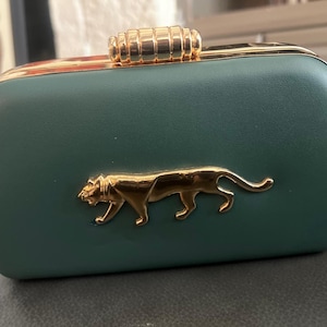 May include: A rectangular, forest green clutch with a gold-colored clasp and trim. A gold-colored panther emblem is centered on the front. The clutch is likely made of a smooth, leather-like material.