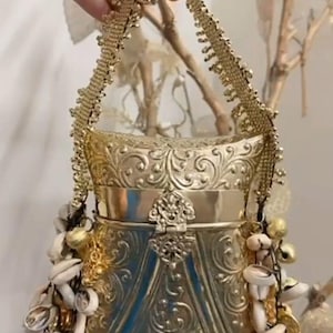 May include: A gold-toned metal handbag featuring an intricate embossed floral pattern across its surface. The bag has a structured frame with a decorative latch closure and a beaded handle. White cowrie shells and small gold bells hang from the sides.