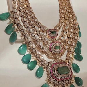 May include: A multi-layered gold necklace featuring three central rectangular pendants set with green stones and pink accents, surrounded by clear crystals. The design includes numerous dangling, teardrop-shaped green beads.